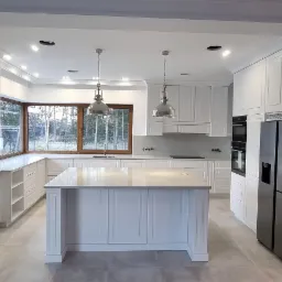 Jasna, przestronna kuchnia w stylu klasycznym z wyspą, białymi frontami szafek z frezowanymi detalami, dużym oknem narożnym i metalowymi lampami wiszącymi.