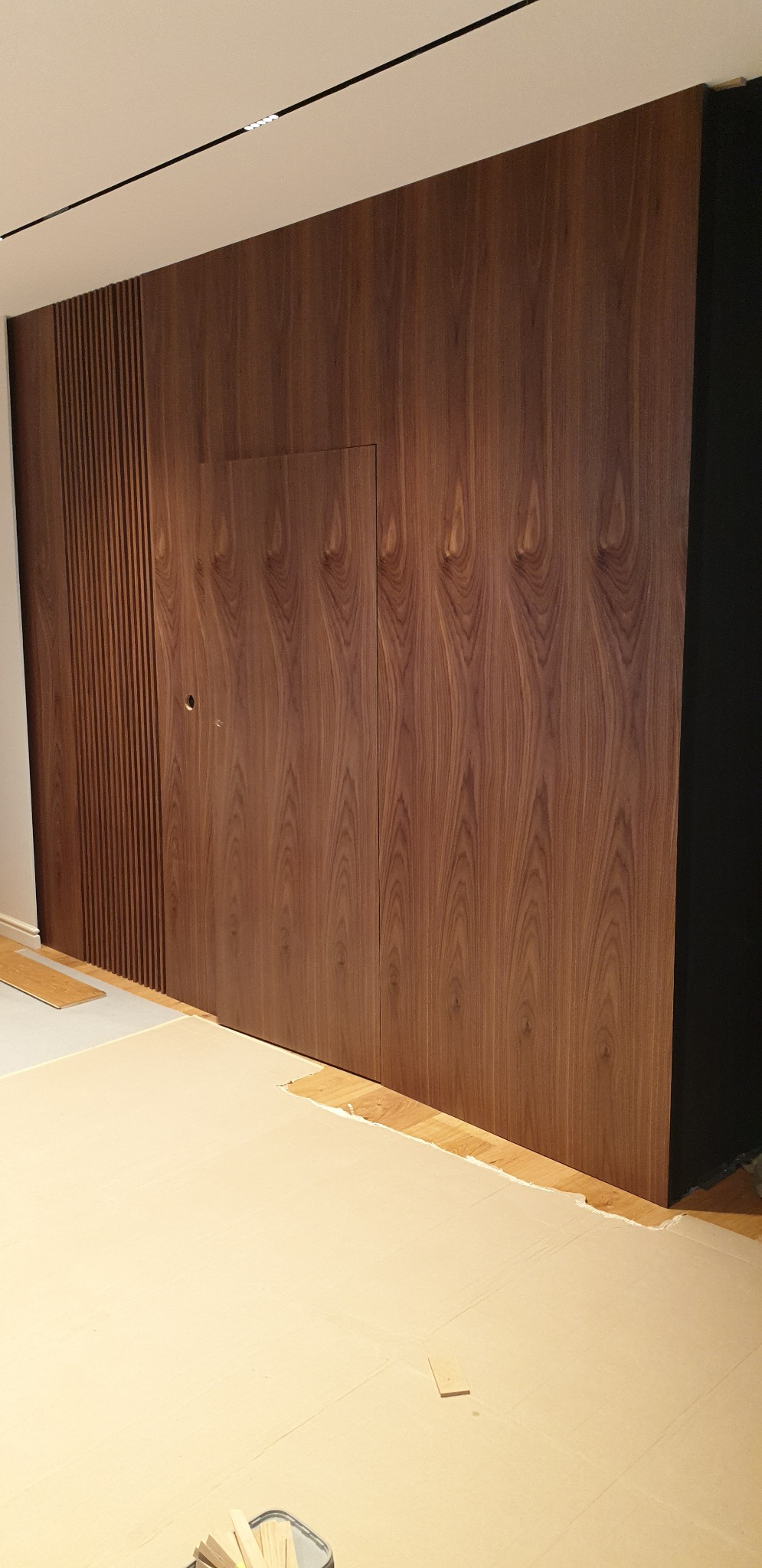 Wbudowana szafa z ukrytymi drzwiczkami, wykończona fornirem z orzecha włoskiego, z pionowymi listwami po lewej stronie, podczas montażu, zabezpieczona kartonem na podłodze.
