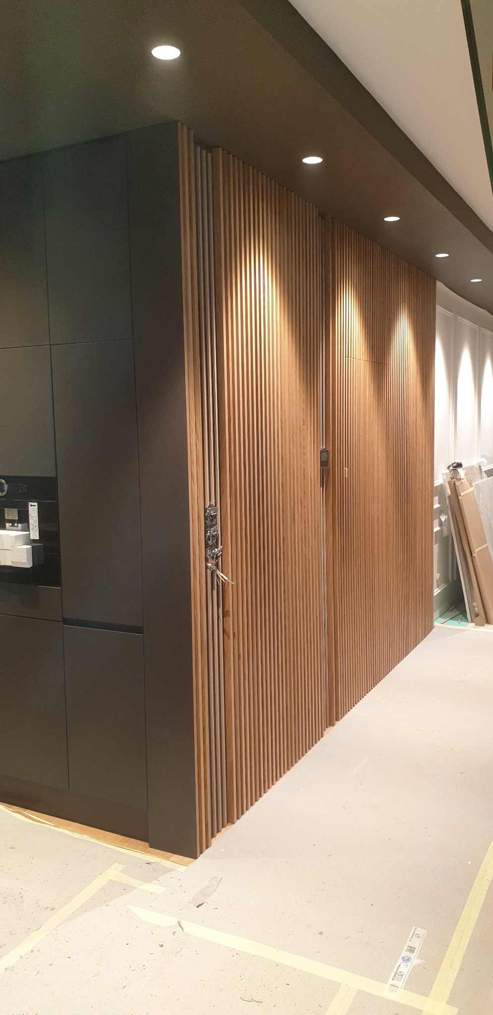 Ciemna zabudowa kuchenna sąsiaduje z drewnianą ścianą z pionowych listewek, widoczne prace wykończeniowe.