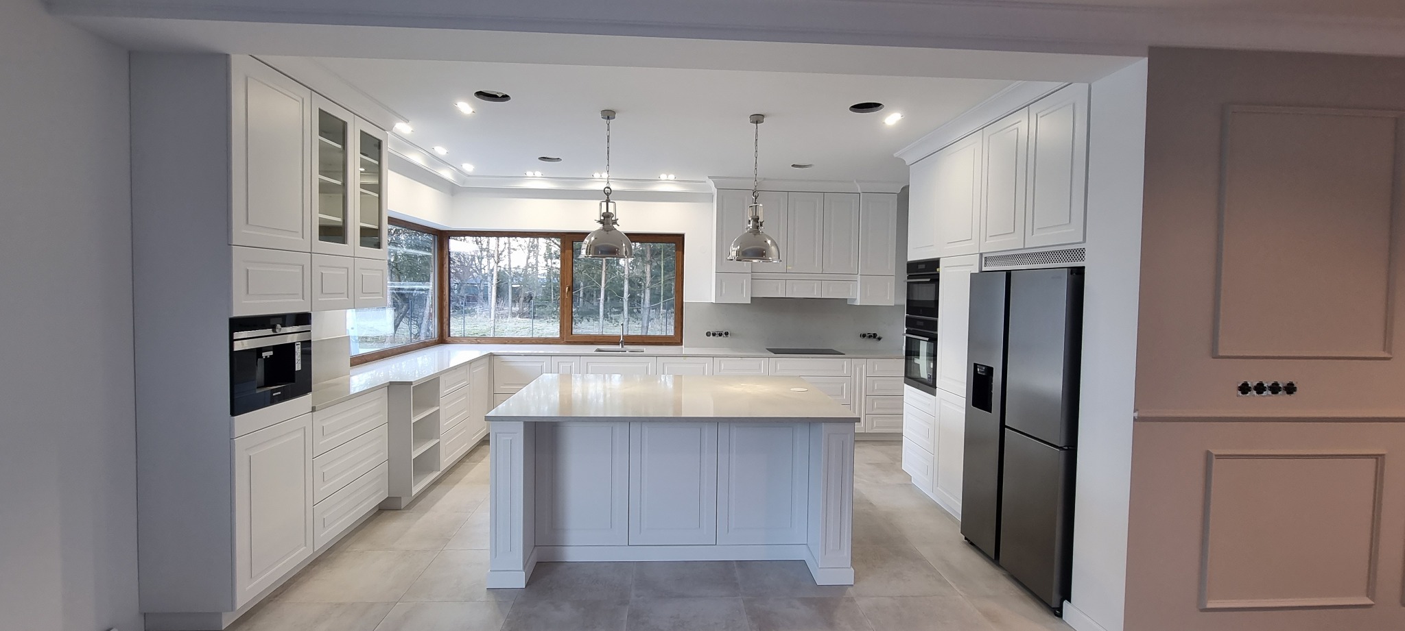 Jasna, przestronna kuchnia w stylu klasycznym z wyspą, białymi frontami szafek z frezowanymi detalami, dużym oknem narożnym i metalowymi lampami wiszącymi.