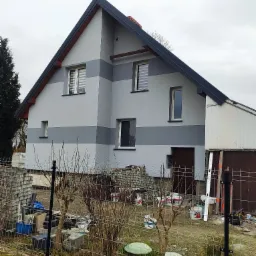 Dwukondygnacyjny dom z szarą elewacją w pasy, widoczne puszki z farbą i materiały budowlane na posesji za ogrodzeniem.