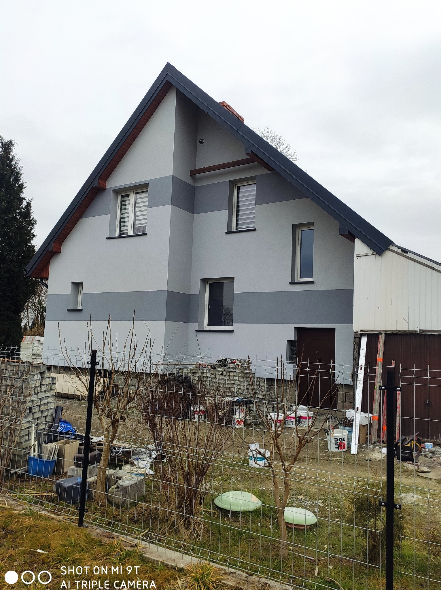 Dwukondygnacyjny dom z szarą elewacją w pasy, widoczne puszki z farbą i materiały budowlane na posesji za ogrodzeniem.