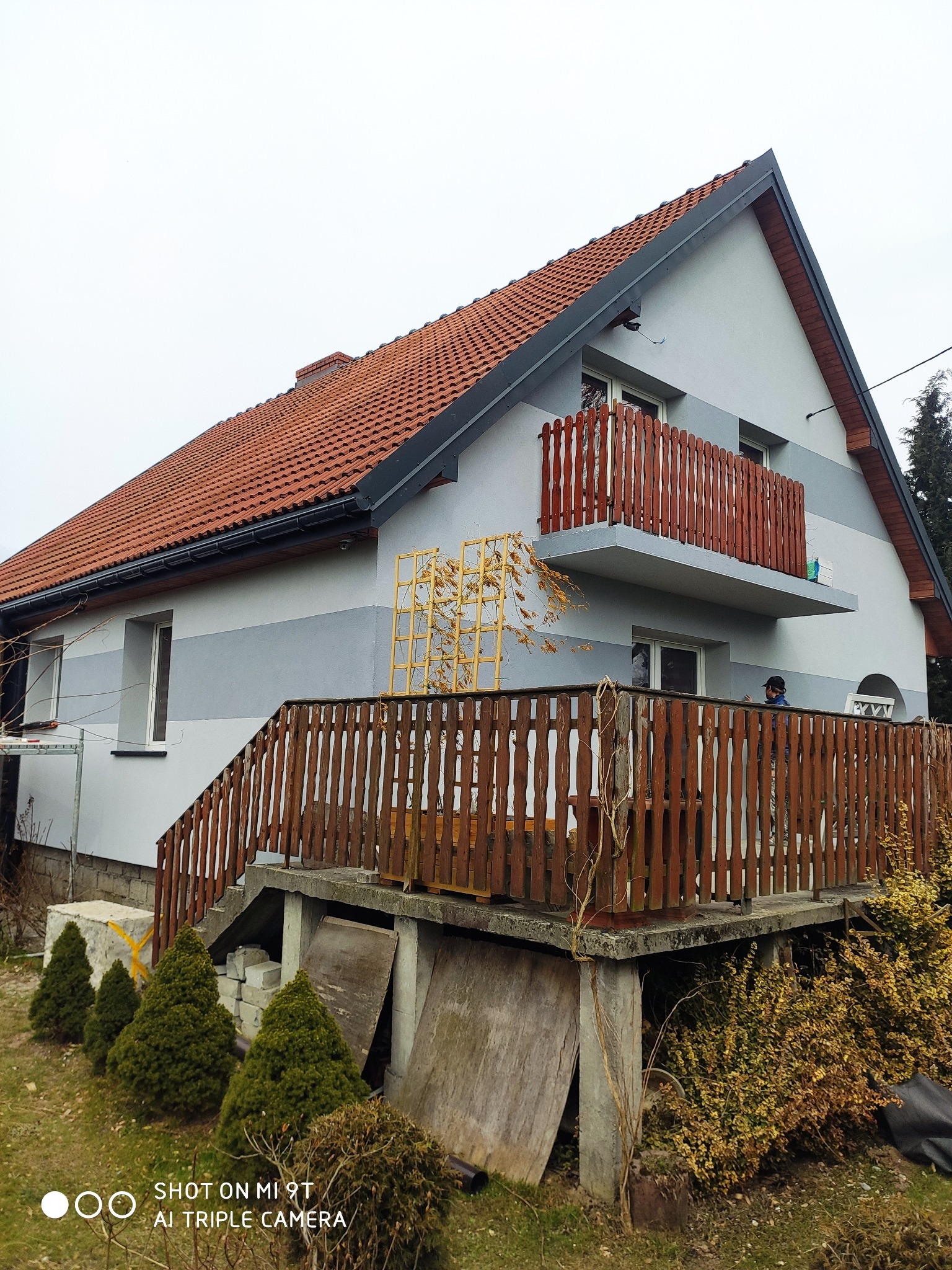 Dwukondygnacyjny dom z czerwonym dachem i szarą elewacją z drewnianą balustradą balkonu i werandy, widok z dołu, pochmurne niebo.