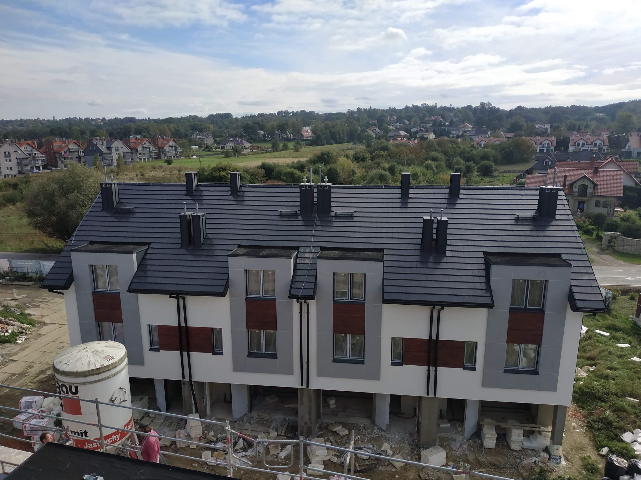 Nowoczesny szereg domów w trakcie budowy, widoczne rusztowania, materiały budowlane i zbiornik na zaprawę, w tle zabudowa jednorodzinna i zieleń.