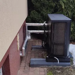 Czarna pompa ciepła zamontowana na zewnątrz budynku, z izolowanymi rurami prowadzącymi wzdłuż ściany. Widoczne tuje w tle.