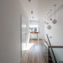 Nowoczesny, minimalistyczny korytarz z drewnianą podłogą, białymi ścianami i szklaną balustradą schodów. Sufit zdobią kuliste lampy wiszące i punktowe reflektory. W głębi widoczne okno z roślinami...