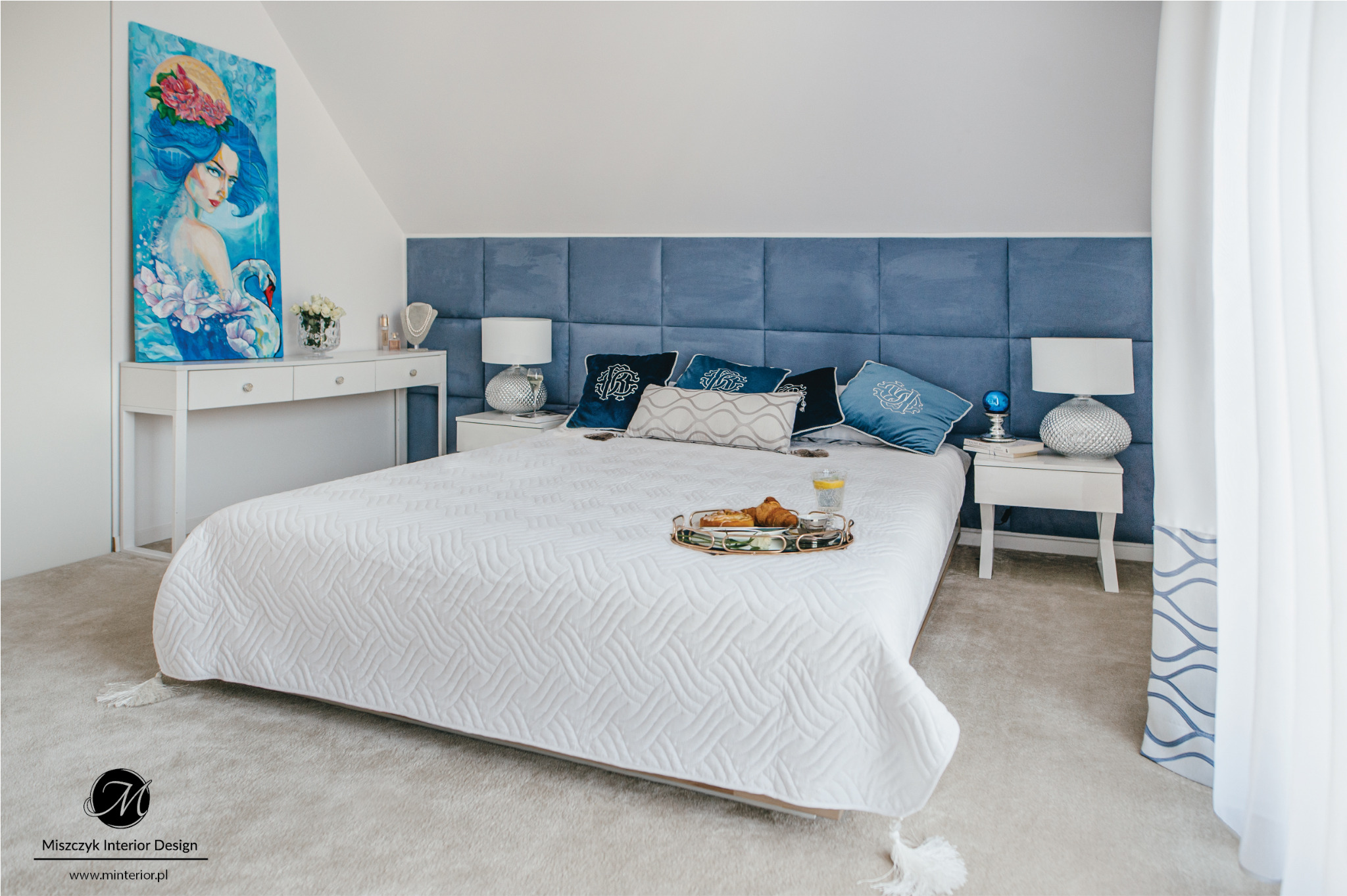 Jasna sypialnia w Katowicach z niebieskim zagłówkiem i dekoracyjnymi poduszkami, na łóżku taca ze śniadaniem, nad komodą obraz z motywem kobiety i łabędzia.