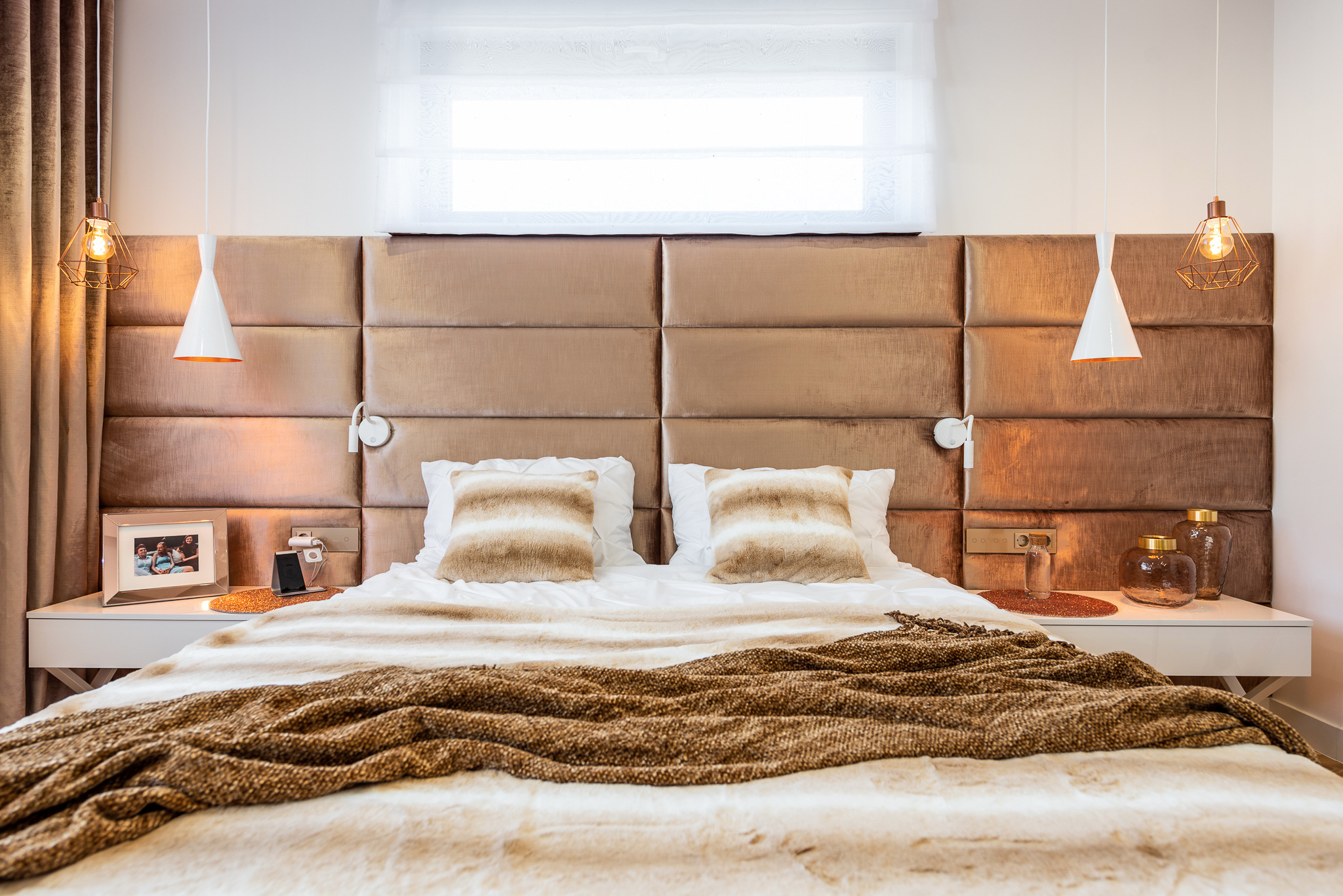 Sypialnia z tapicerowanym panelem ściennym w kolorze taupe, białymi szafkami nocnymi i dekoracyjnymi lampami wiszącymi o stożkowym kształcie, uzupełniona o tekstylia w odcieniach brązu i beżu.