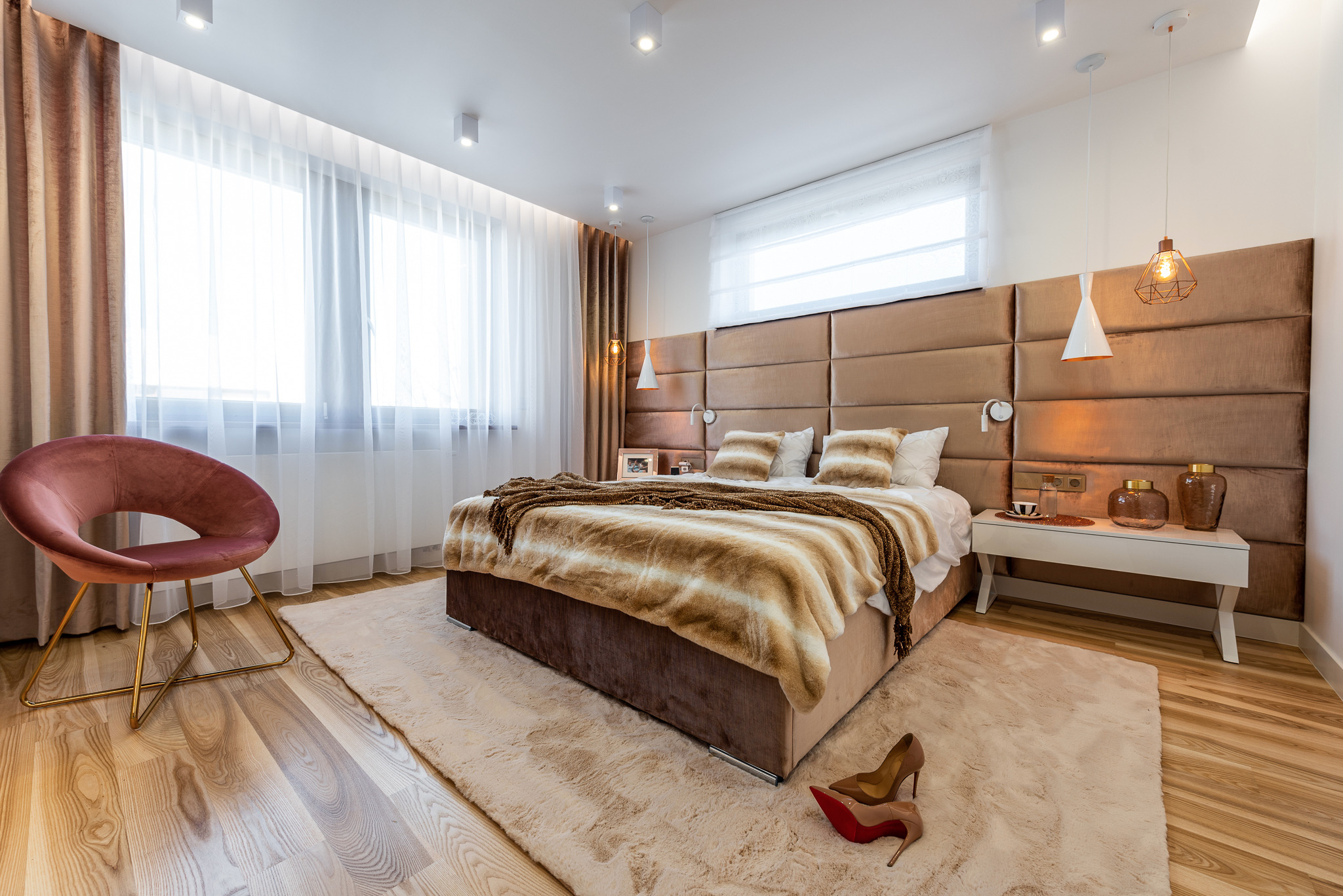 Nowoczesna sypialnia z dużym tapicerowanym zagłówkiem w kolorze brązu, puszystym dywanem i krzesłem w stylu retro. Na podłodze leżą damskie szpilki z czerwoną podeszwą.