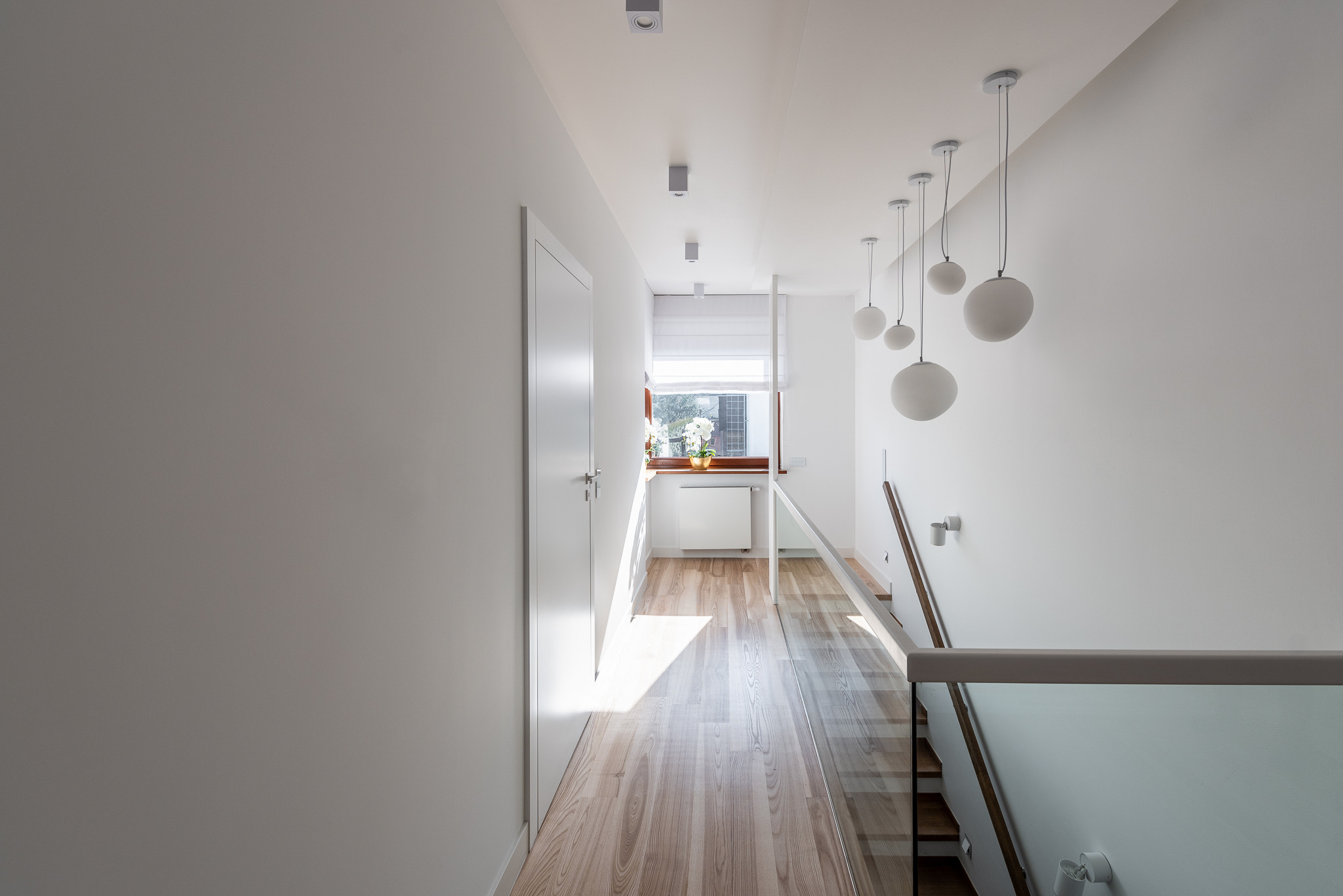 Nowoczesny, minimalistyczny korytarz z drewnianą podłogą, białymi ścianami i szklaną balustradą schodów. Sufit zdobią kuliste lampy wiszące i punktowe reflektory. W głębi widoczne okno z roślinami...