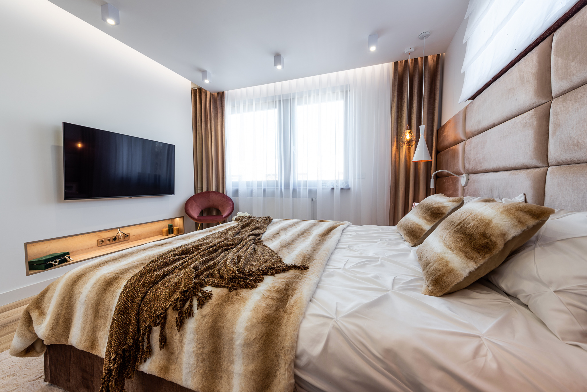 Nowoczesna sypialnia z tapicerowanym zagłówkiem, telewizorem na ścianie i dekoracyjną niszą z oświetleniem, uzupełniona futrzaną narzutą i poduszkami.