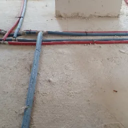 Wnętrze w trakcie remontu z widocznymi czerwonymi i niebieskimi rurami instalacji rozprowadzonymi po podłodze, zabezpieczonymi folią, oraz fragmentami ścian w stanie surowym.