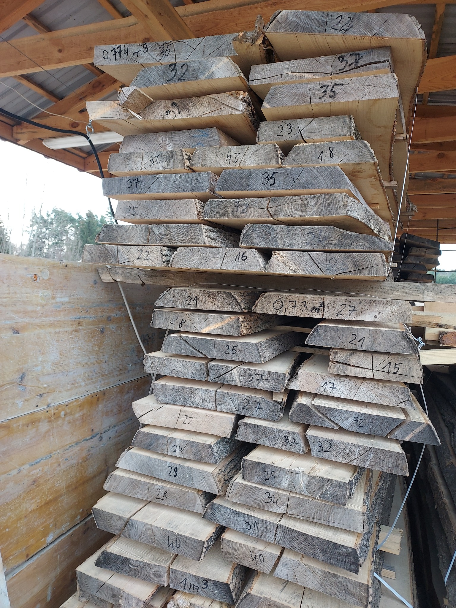 Stos tarcicy ułożonej pod zadaszeniem; deski z napisanymi markerem numerami i oznaczeniami objętości.