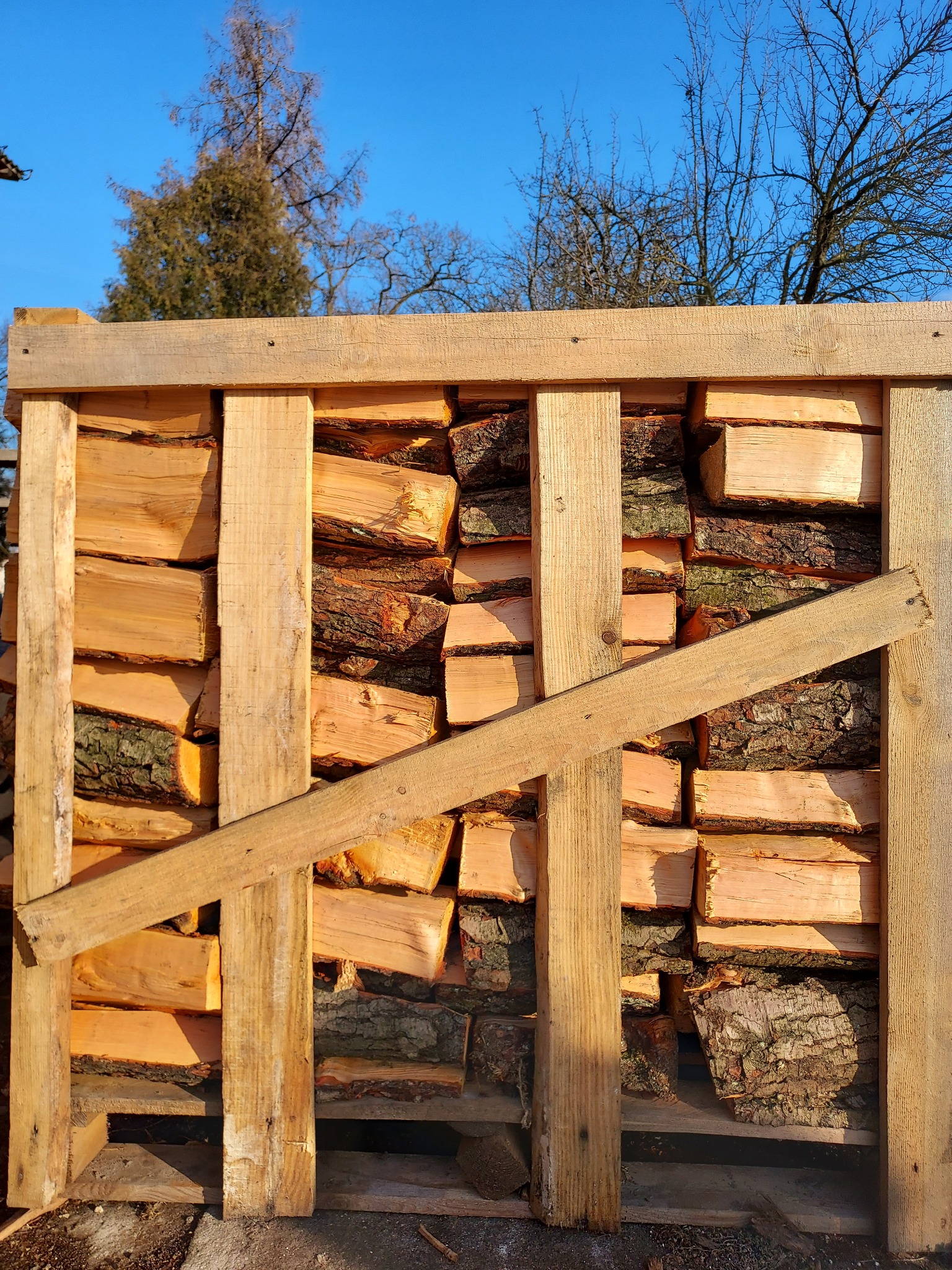 Równomiernie ułożone, pocięte kawałki drewna w drewnianej skrzyni, na tle błękitnego nieba i drzew bez liści.