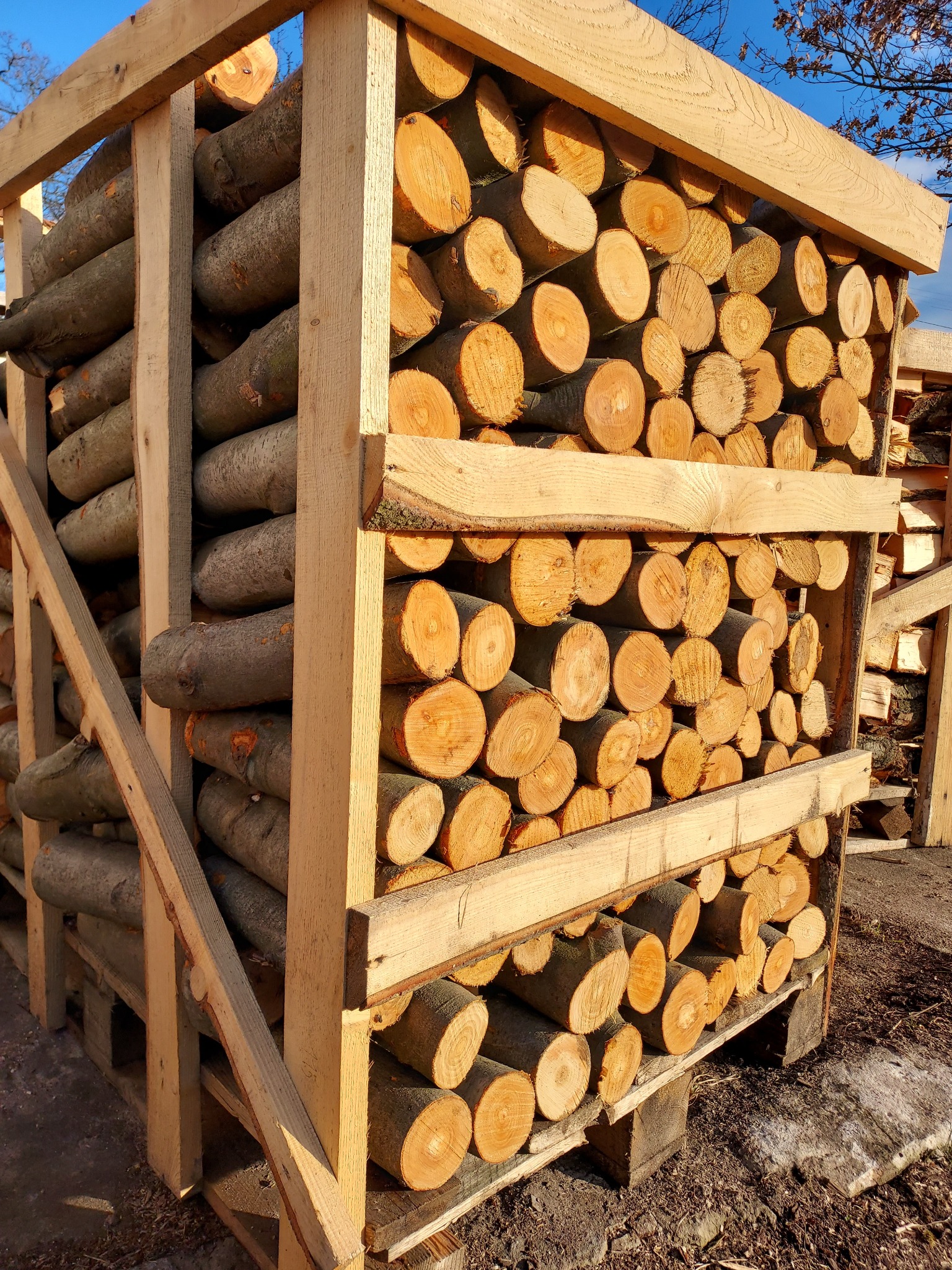 Równomiernie ułożone szczapy drewna w drewnianej skrzyni, widoczne słoje, część z korą, na zewnątrz w słoneczny dzień.