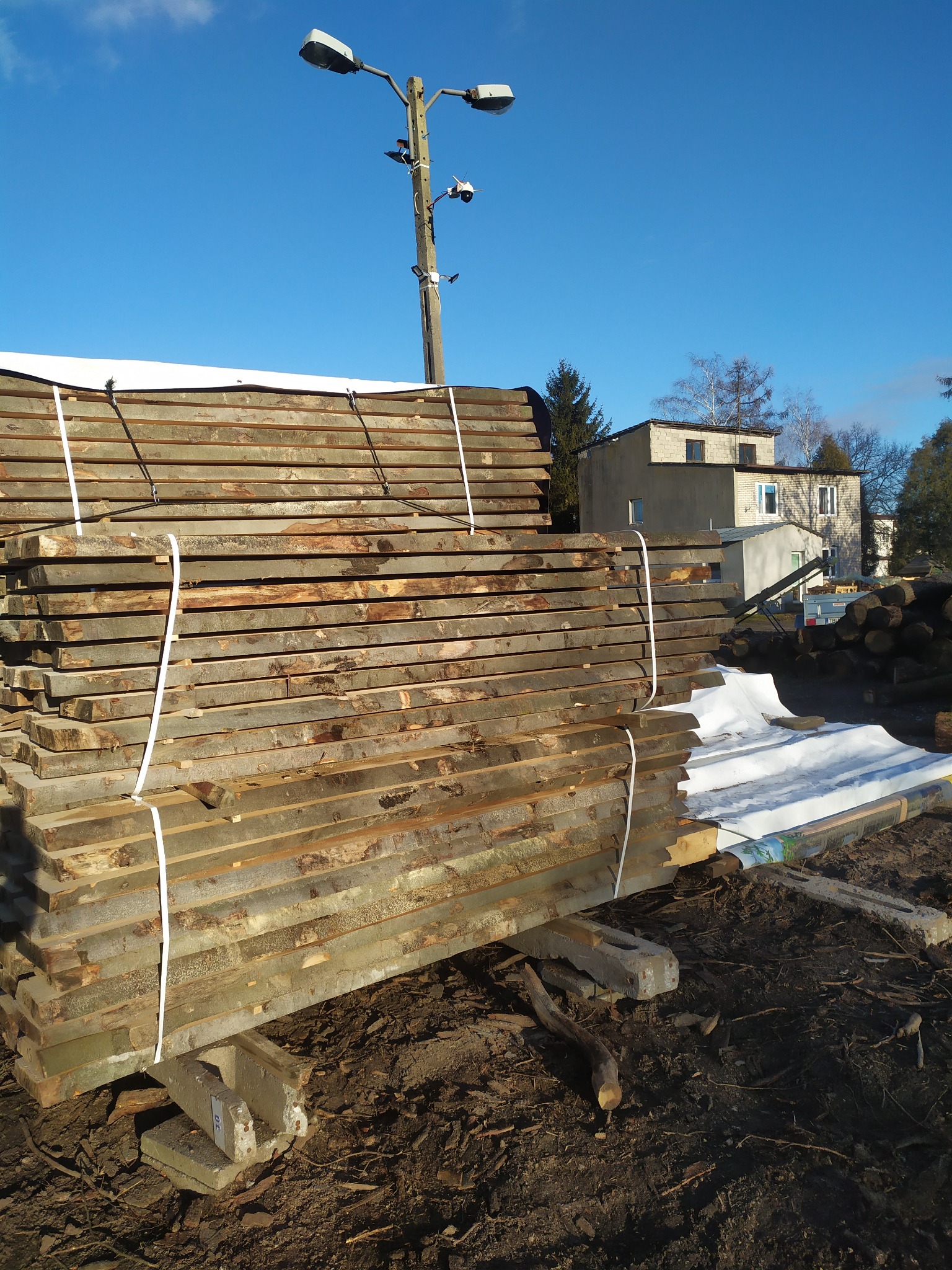 Stos surowej tarcicy budowlanej zabezpieczony białym sznurkiem, ułożony na betonowych wspornikach na ziemi, w tle budynki i słup oświetleniowy pod błękitnym niebem.