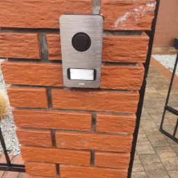 Szczegółowe ujęcie domofonu Urmet z metalową obudową, zamontowanego na ceglanym słupku ogrodzeniowym z widocznymi śladami białej farby.