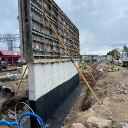 Wysoki betonowy fundament z deskowaniem i hydroizolacją w wykopie, widoczne niebieskie rury, w tle plac budowy z dźwigiem i wagonem kolejowym.