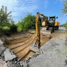 Żółta koparka Caterpillar pogłębia wykop w asfalcie, obok stoi wywrotka, tło stanowią drzewa i błękitne niebo z chmurami