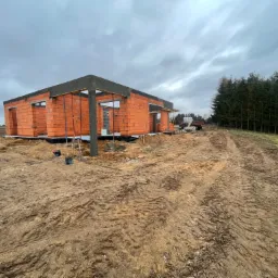 Surowy stan domu jednorodzinnego z czerwonej cegły, widoczne szalunki stropowe, teren wokół w stanie naturalnym, błotnisty, pochmurne niebo.