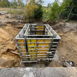 Szare szalunki fundamentowe Doka z żółtymi panelami w wykopie, widoczne zbrojenie i otoczenie budowy z ziemią i roślinnością.