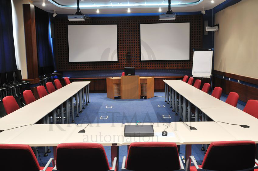 Sala konferencyjna z dwoma ekranami projekcyjnymi, stołami w kształcie podkowy i mównicą, oświetlona lampami sufitowymi i projektorami.