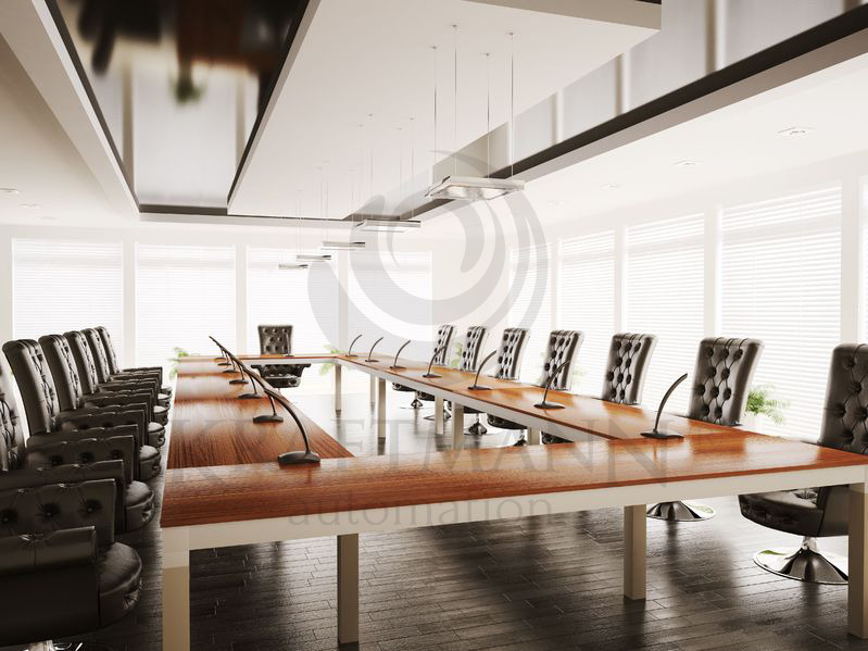 Sala konferencyjna z długim drewnianym stołem, czarnymi skórzanymi krzesłami z guzikami i mikrofonami, oświetlona wiszącymi lampami i naturalnym światłem z okien.