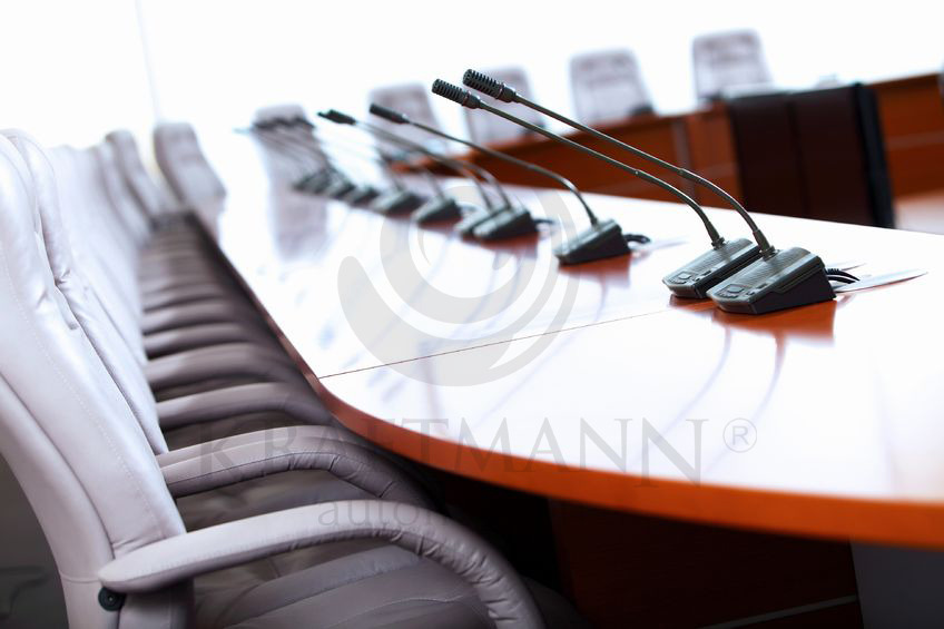 Sala konferencyjna z długim stołem i rzędem mikrofonów na gęsich szyjach, ustawionych przed pustymi, skórzanymi krzesłami.