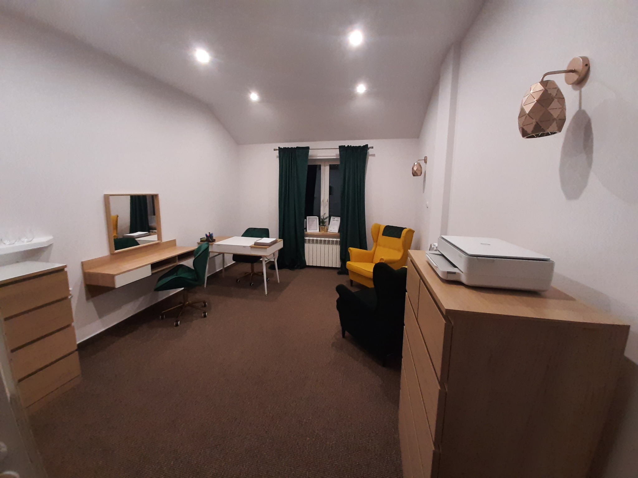 Jasny gabinet z biurkiem, krzesłami w odcieniach zieleni, żółtym fotelem, komodami i drukarką. Okno z ciemnozielonymi zasłonami i dekoracyjnymi lampami ściennymi.