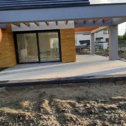 Nowoczesny taras z jasnoszarej kostki brukowej ułożonej przy domu z drewnianą elewacją, widoczne czarne obrzeża i przygotowany teren pod ogród.