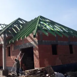 Budowa domu jednorodzinnego w trakcie wznoszenia, widoczna konstrukcja z czerwonej cegły i zielona więźba dachowa, na pierwszym planie palety z materiałami budowlanymi i pracownik z drabiną.