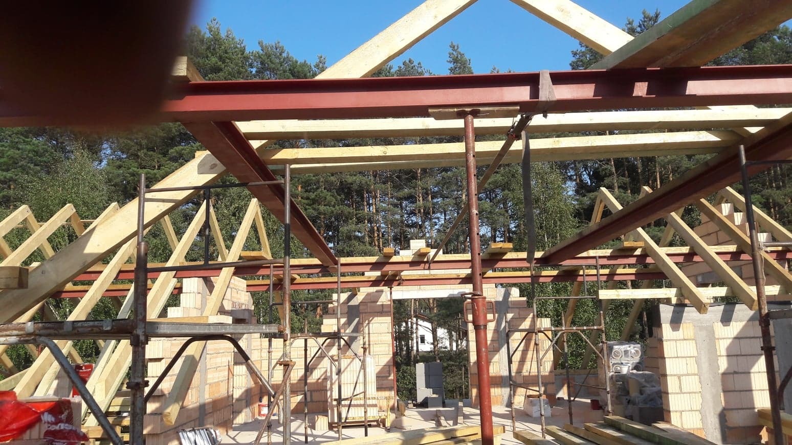Konstrukcja dachu w trakcie budowy, widoczne drewniane elementy więźby dachowej, stalowe belki, murowane ściany budynku w stanie surowym otwartym, las w tle.