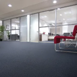 Biuro z ciemną, teksturowaną podłogą, szklanymi ścianami działowymi, widocznymi przestrzeniami roboczymi i czerwonymi krzesłami w tle.