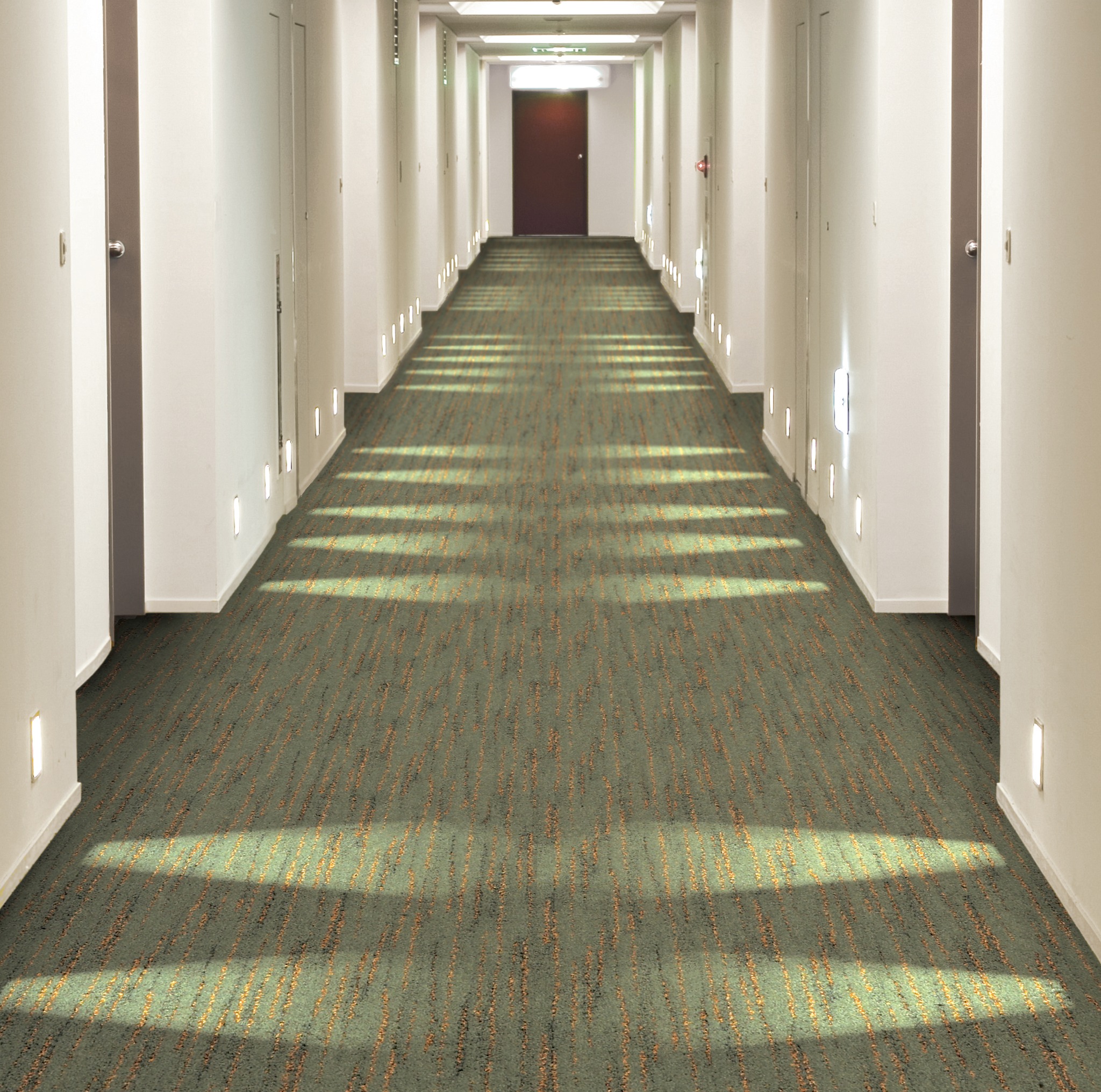 Długi korytarz hotelowy z zieloną wykładziną w paski i symetrycznie rozmieszczonymi drzwiami oraz oświetleniem przypodłogowym.