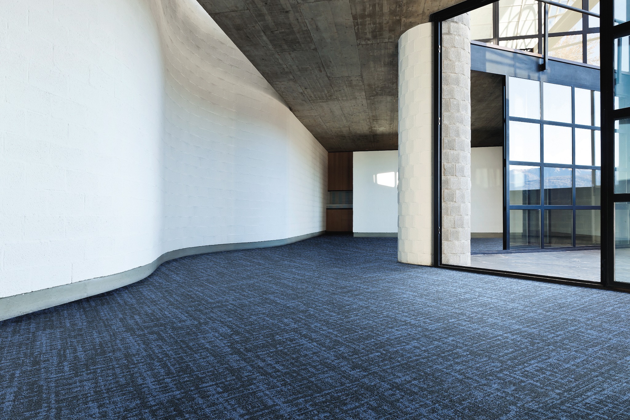 Przestronne wnętrze z ciemnoniebieską wykładziną w geometryczny wzór, białe, zaokrąglone ściany z cegły, surowy betonowy sufit i duże, przeszklone drzwi przesuwne.