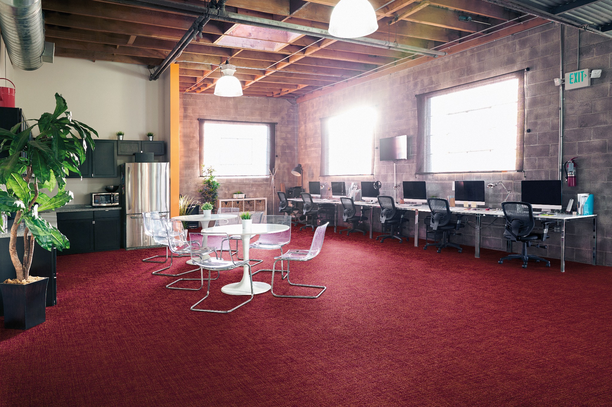 Biuro z bordową tekstylną podłogą, białymi okrągłymi stołami z przezroczystymi krzesłami i rzędami biurek komputerowych w tle.