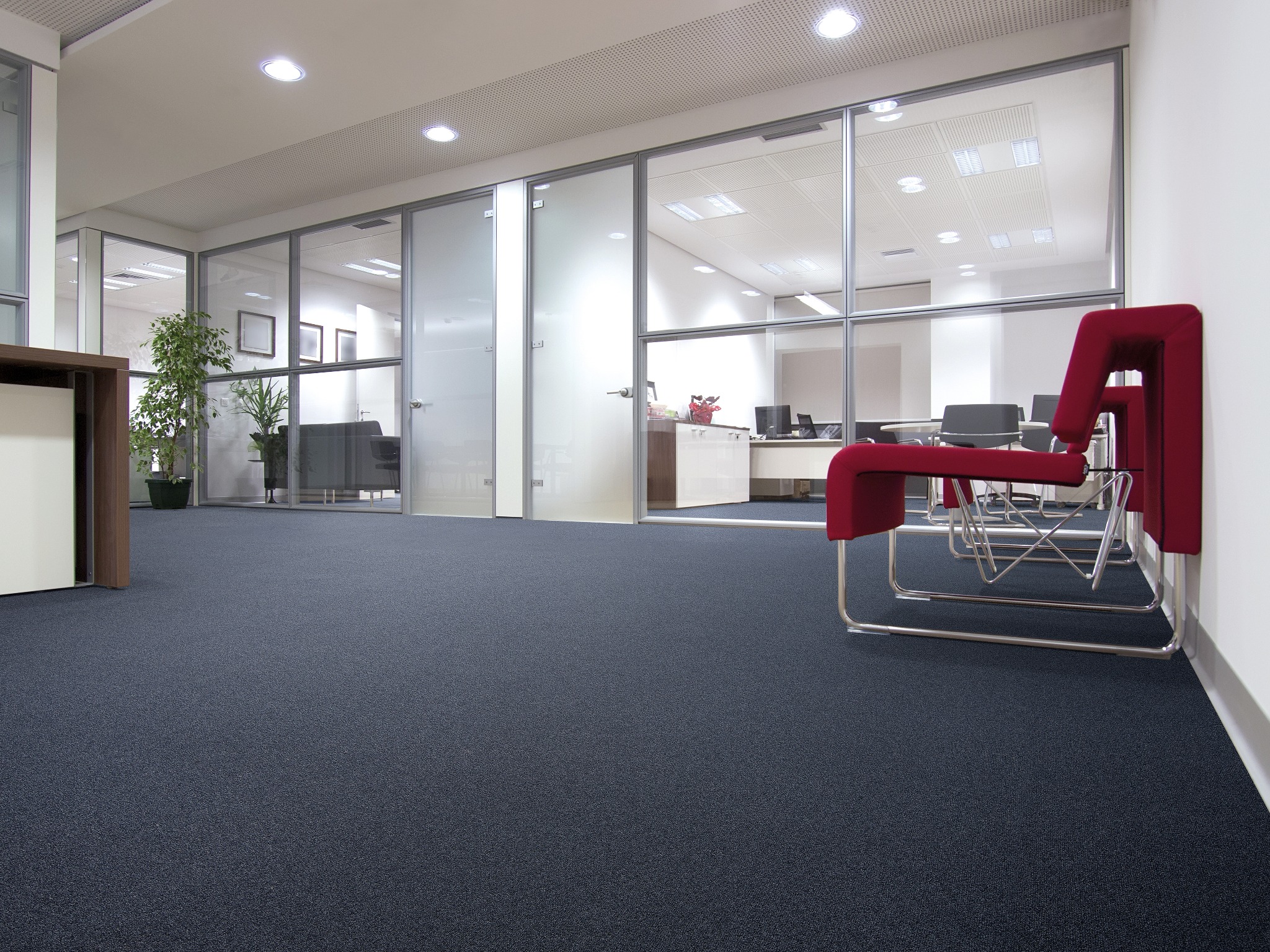 Biuro z ciemną, teksturowaną podłogą, szklanymi ścianami działowymi, widocznymi przestrzeniami roboczymi i czerwonymi krzesłami w tle.