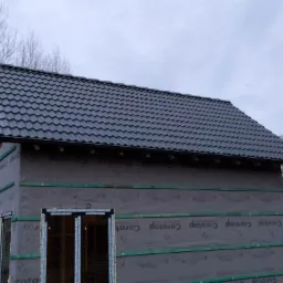 Nowy, grafitowy dach z blachodachówki modułowej i zamontowane rynny na budynku w trakcie ocieplania, widoczna membrana dachowa i elementy konstrukcji drewnianej.