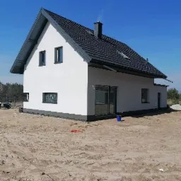 Nowoczesny dom jednorodzinny z białą elewacją, ciemną dachówką i szarymi detalami, widoczny z boku na tle błękitnego nieba, z niezagospodarowanym terenem wokół.