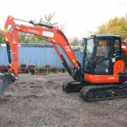 Pomarańczowa minikoparka Kubota KX057-4 na nierównym terenie, z wyeksponowaną łyżką i dodatkowymi łyżkami w tle, obok kontenera i drzew.