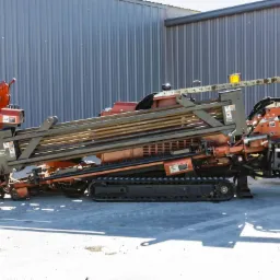 Pomarańczowa wiertnica kierunkowa Ditch Witch JT4020 na gąsienicach, z widocznymi rzędami wierteł i metalową konstrukcją, na tle szarego magazynu.