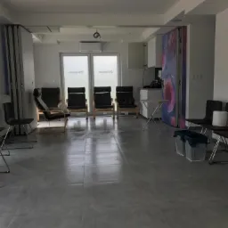 Przestronne, jasne wnętrze biurowe z szarymi płytkami podłogowymi, czarnymi krzesłami i fotelami, białymi ścianami i sufitem, widocznymi elementami instalacji oraz ścianą z grafiką. Widoczne kosze...