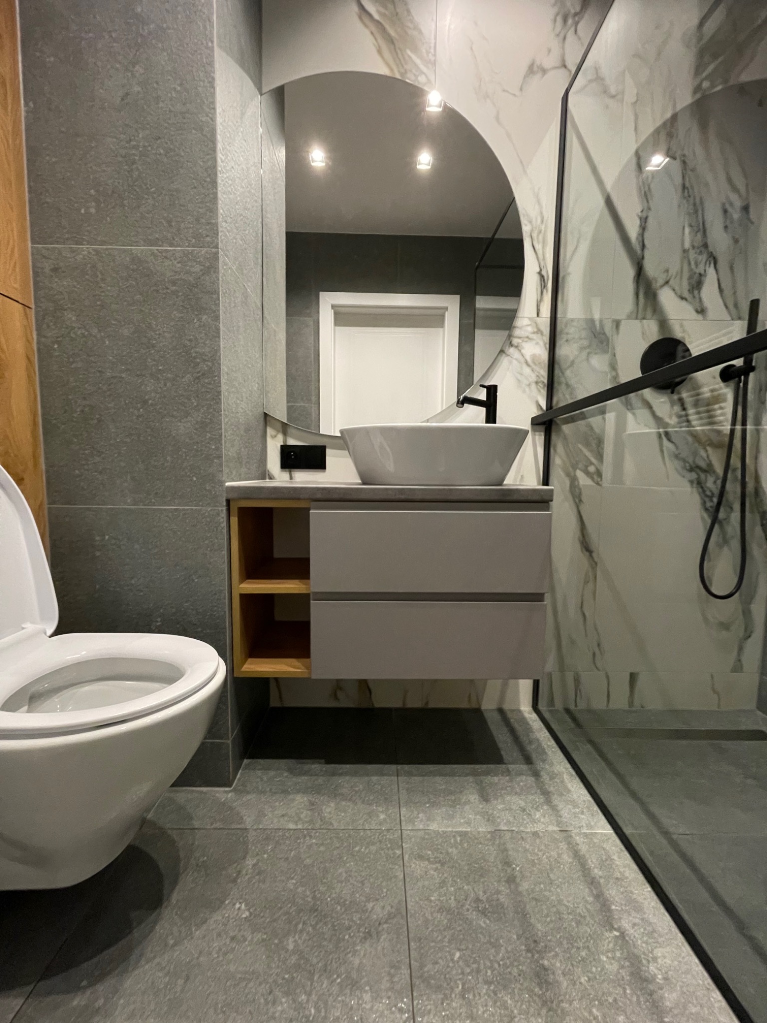 Nowoczesna łazienka z szarą płytką, umywalką nablatową na szafce z drewnianymi półkami, okrągłym lustrem i kabiną prysznicową ze szklaną ścianą.