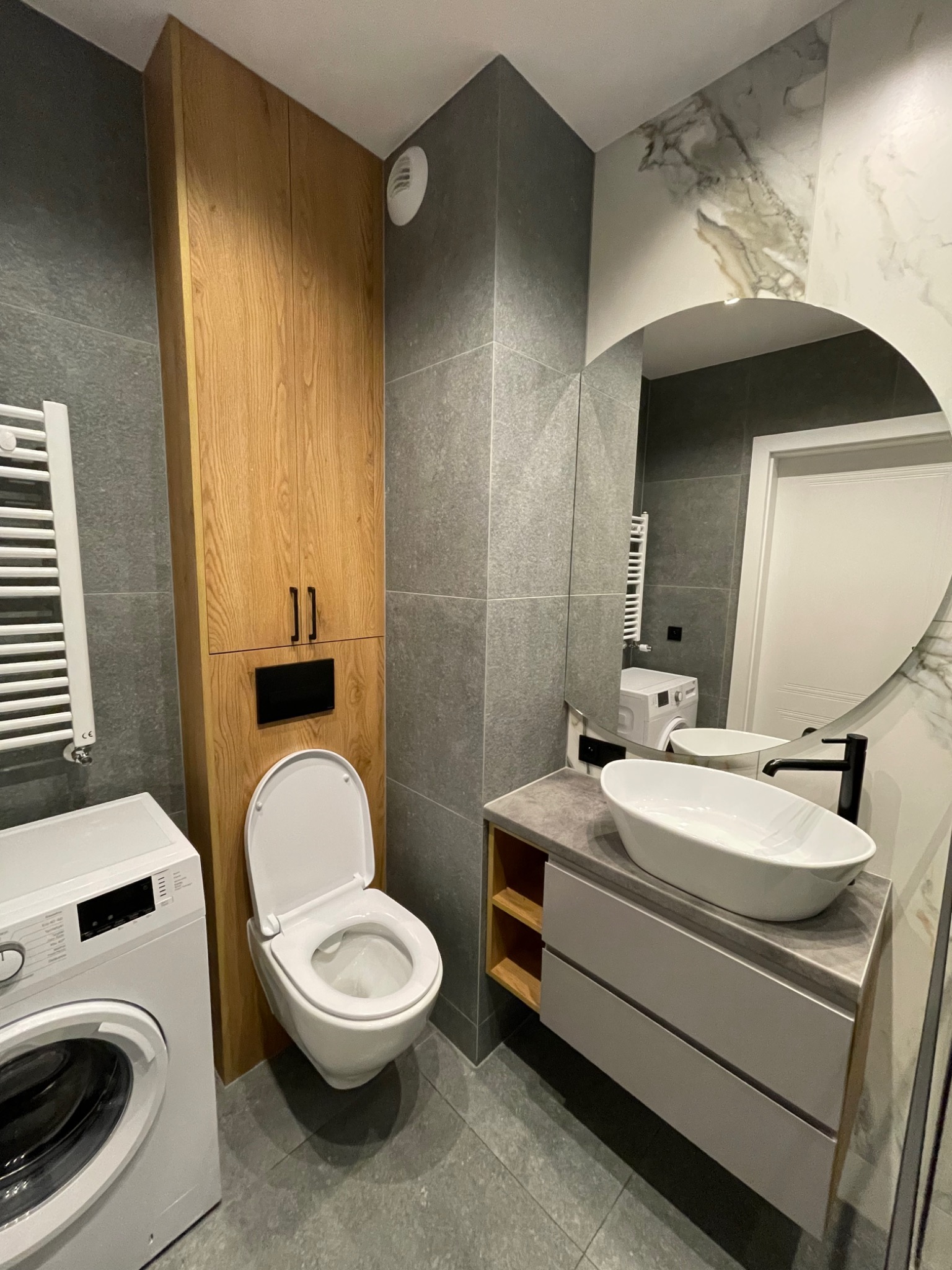 Nowoczesna łazienka z szarymi płytkami, podwieszaną toaletą, drewnianą szafką, umywalką nablatową i okrągłym lustrem odbijającym pralkę.