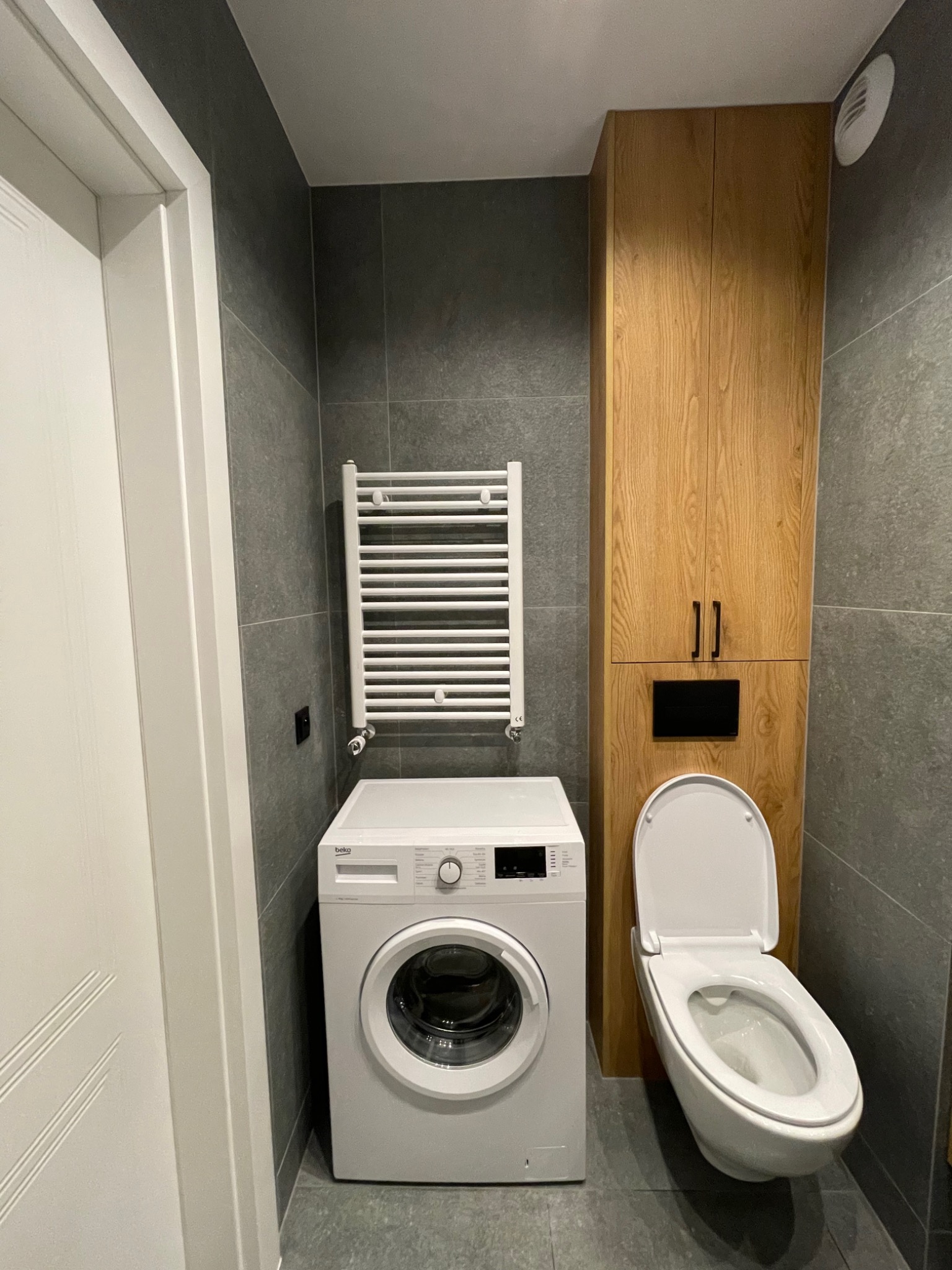 Nowoczesna łazienka z szarymi płytkami, drewnianą szafką, białą pralką Beko i grzejnikiem drabinkowym, toaleta z deską wolnoopadającą.