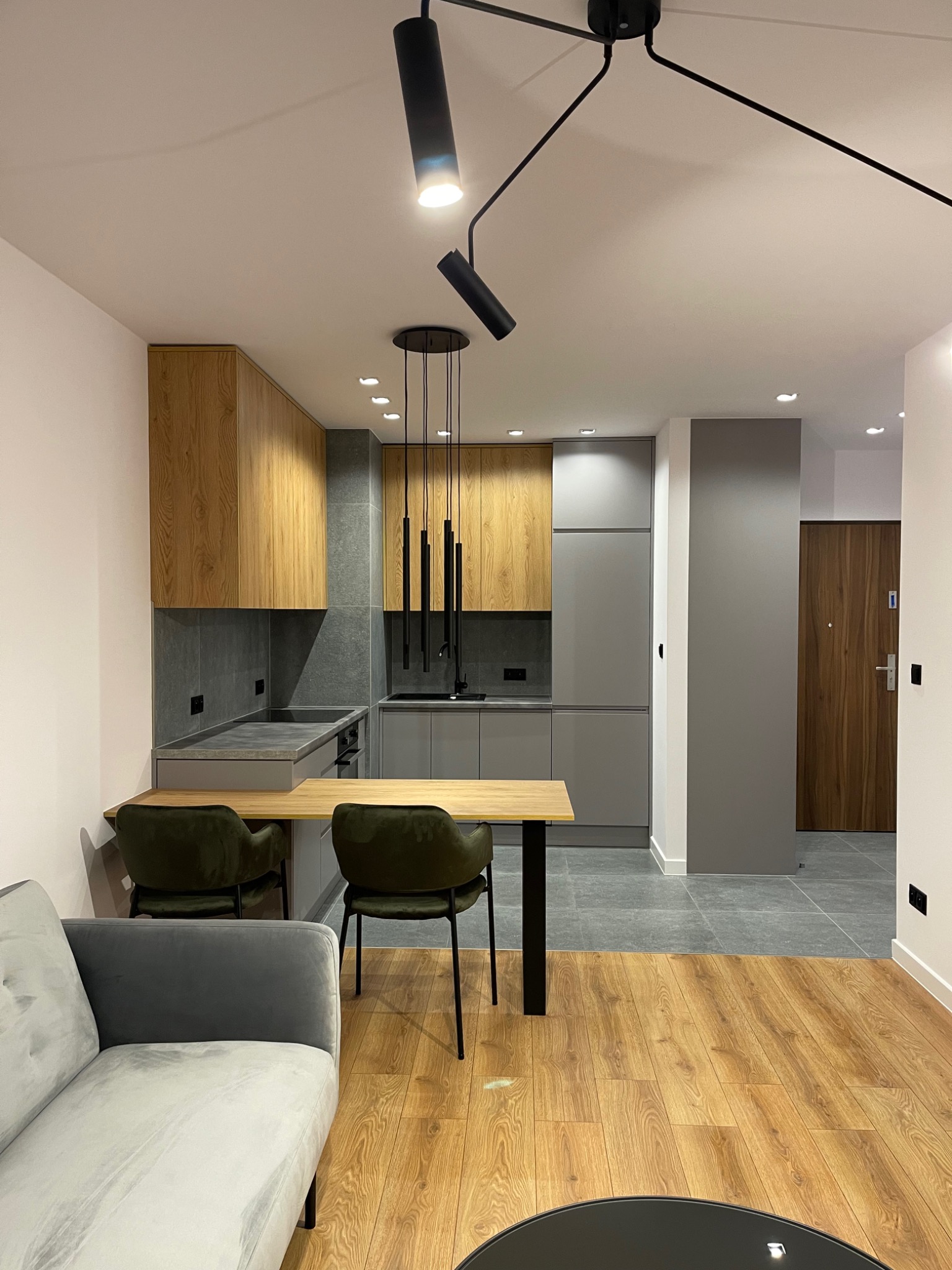 Nowoczesne wnętrze: kuchnia z drewnianymi i szarymi szafkami, szary blat, stół z krzesłami, sofa i drewniana podłoga, oświetlenie punktowe i wiszące lampy.