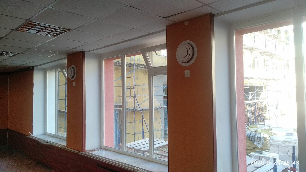 Wnętrze biurowe z widocznymi nawiewnikami sufitowymi na ścianach koloru brązowego, okna z widokiem na rusztowania i materiały budowlane na zewnątrz.