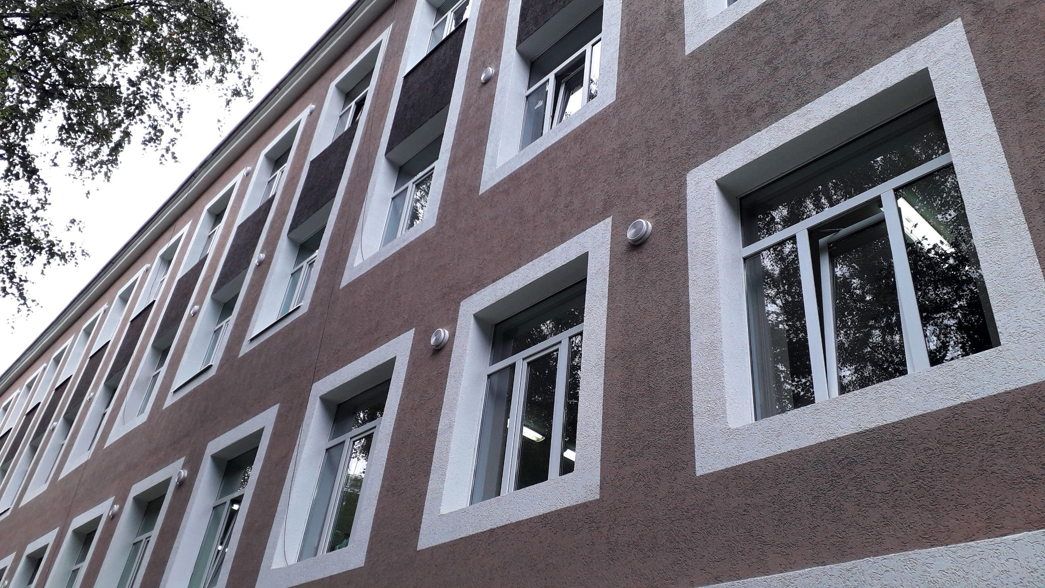 Elewacja budynku z oknami wentylacyjnymi, widoczne kratki wentylacyjne nad niektórymi oknami, brązowy tynk strukturalny i białe obramowania okien.