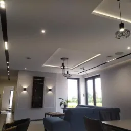 Nowoczesny salon z podwieszanym sufitem, oświetleniem punktowym i dekoracyjnymi lampami wiszącymi, szara sofa i ściany.