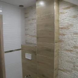 Nowoczesna łazienka z kombinacją białych, poziomych płytek, paneli imitujących drewno i kamiennej okładziny ściany w odcieniach beżu i szarości, z widocznym przyciskiem spłuczki podtynkowej.